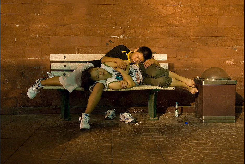 A teenage couple sleeps on a bench using each another as cushions.